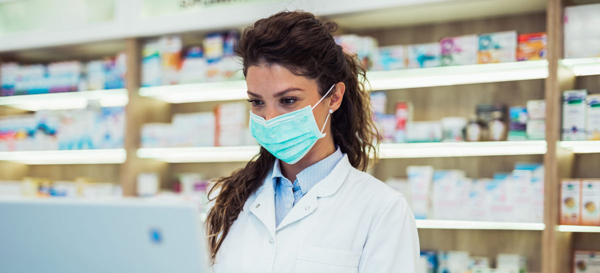 Pharmacist with brunette hair wearing a mask while using a computer in a pharmacy setting